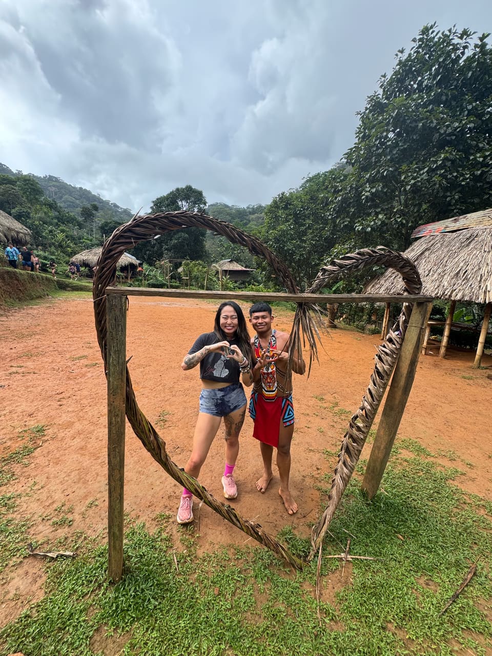 The author and an Embera man make hand-hearts inside a heart-shaped arch of woven branches in a jungle village clearing