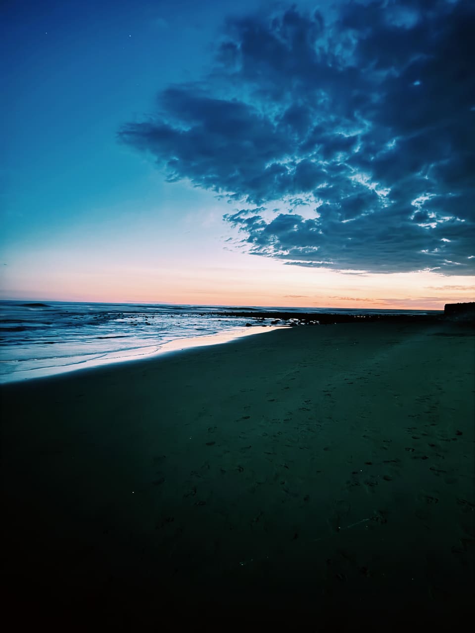 Footprinted dark sand beach at dusk in El Salvador, waves rolling in under heavy clouds and an orange sunset horizon