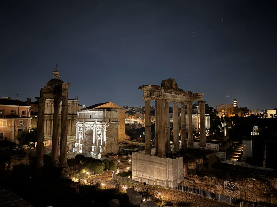 The Roman Forum at night: floodlit temple columns, the Arch of Septimius Severus, and the Colosseum glowing in the distance