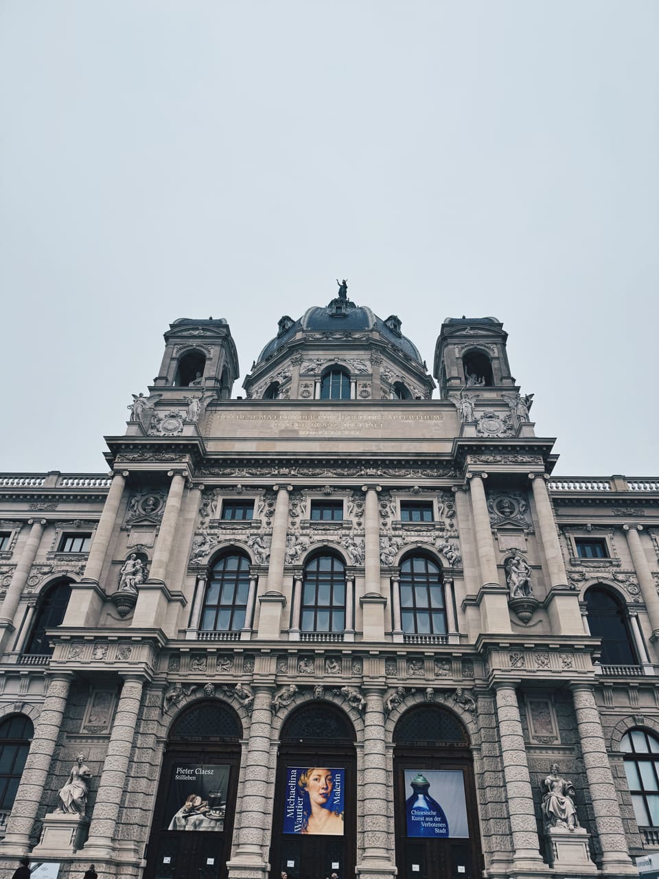 Domed stone facade of Vienna's Kunsthistorisches Museum under a pale grey sky, exhibition banners above its doors