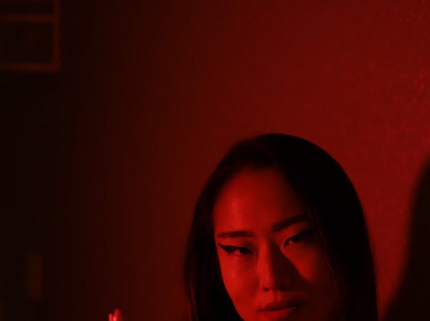 The author in a room lit entirely red, dark hair falling past her shoulders, winged eyeliner, looking down and to the side