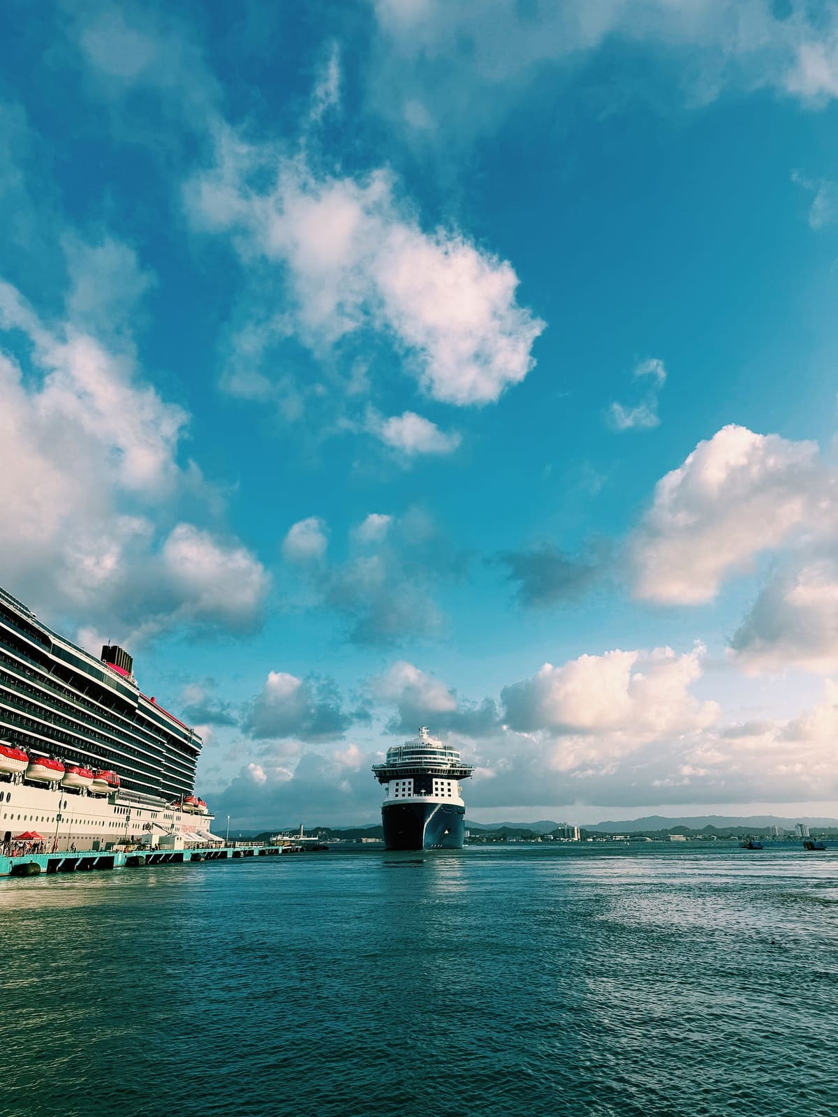 Two cruise ships in a tropical harbor under a sky crowded with cumulus clouds, one docked at a teal pier and one heading out to sea