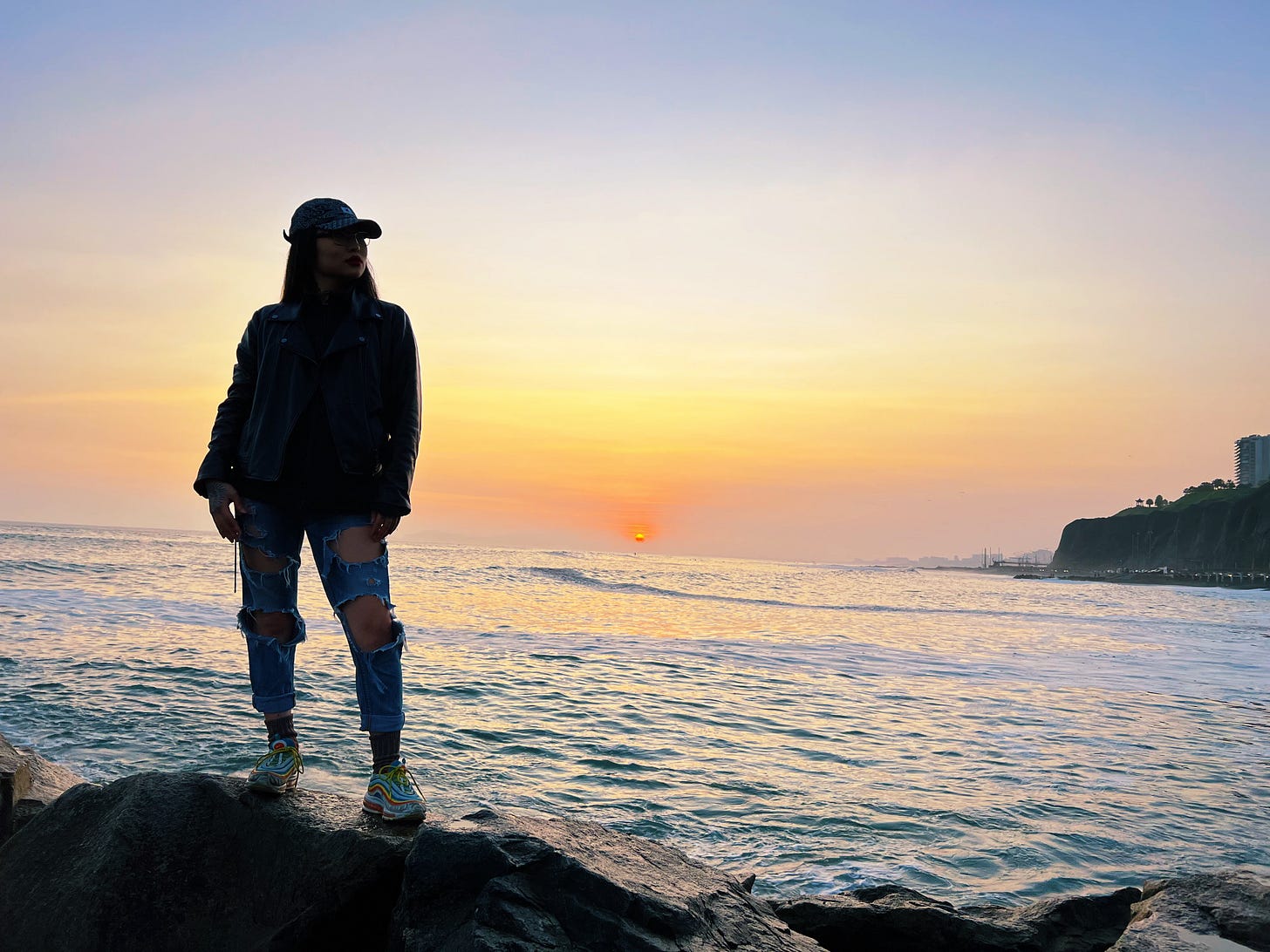 The author in a leather jacket, ripped jeans and a cap stands on a rocky shore at sunset, the sun sinking into the Pacific behind her
