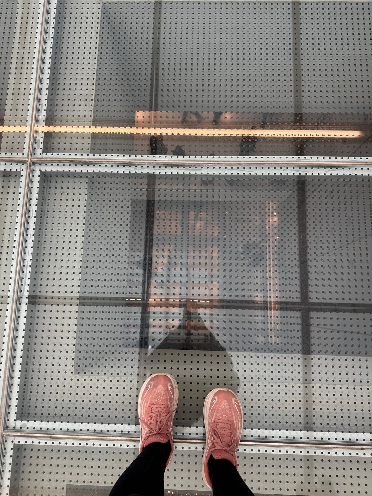 Looking straight down at the author's pink sneakers on a perforated glass floor, the levels of a building visible far below
