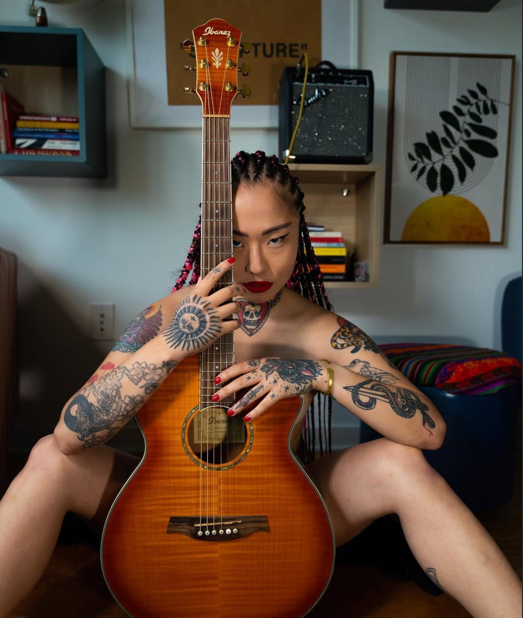The author sits on the floor with an acoustic guitar covering her body, braids threaded with pink, red lipstick and nails, tattooed arms folded over the strings