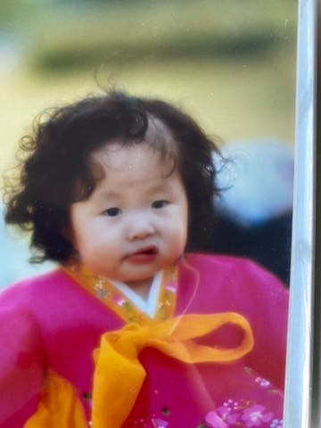 A photographed print of the author as a toddler in a pink hanbok with a yellow bow, dark curls, looking off to one side