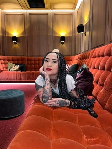 The author lies along an orange velvet banquette in a wood-panelled screening room, chin on her hand, long dark braids, red lipstick, white tee, tattooed arms, a round teal ottoman beside her