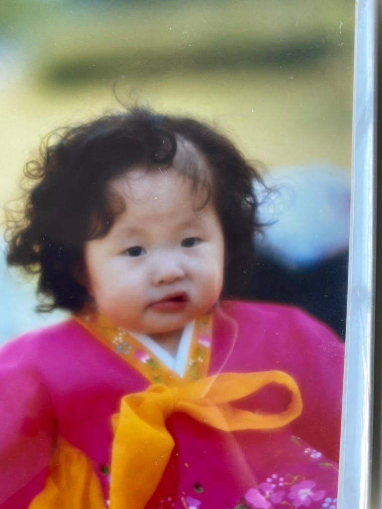 A photographed print of the author as a toddler in a magenta hanbok with an orange-yellow bow, dark curls, looking straight at the camera