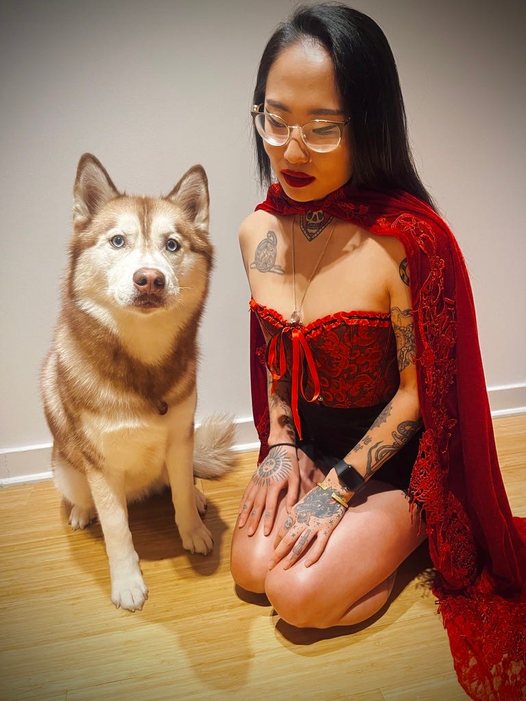 The author kneels on a wood floor beside a red-and-white husky, in a red brocade corset and red lace cape, gold-rimmed glasses, dark red lipstick, eyes lowered