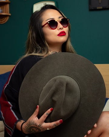 The author in dark sunglasses and red lipstick holds a wide-brimmed olive felt hat in front of her, a tattooed hand on the brim, a teal wall behind