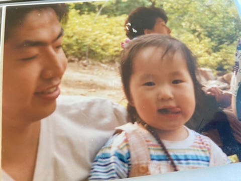 A photographed print of the author as a toddler with a short bob and a striped top, grinning beside a young man in a white shirt who smiles down at her, outdoors among trees