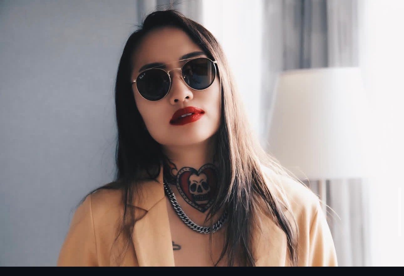 The author in dark round sunglasses and red lipstick, a camel blazer open over the heart and skull tattoo across her throat