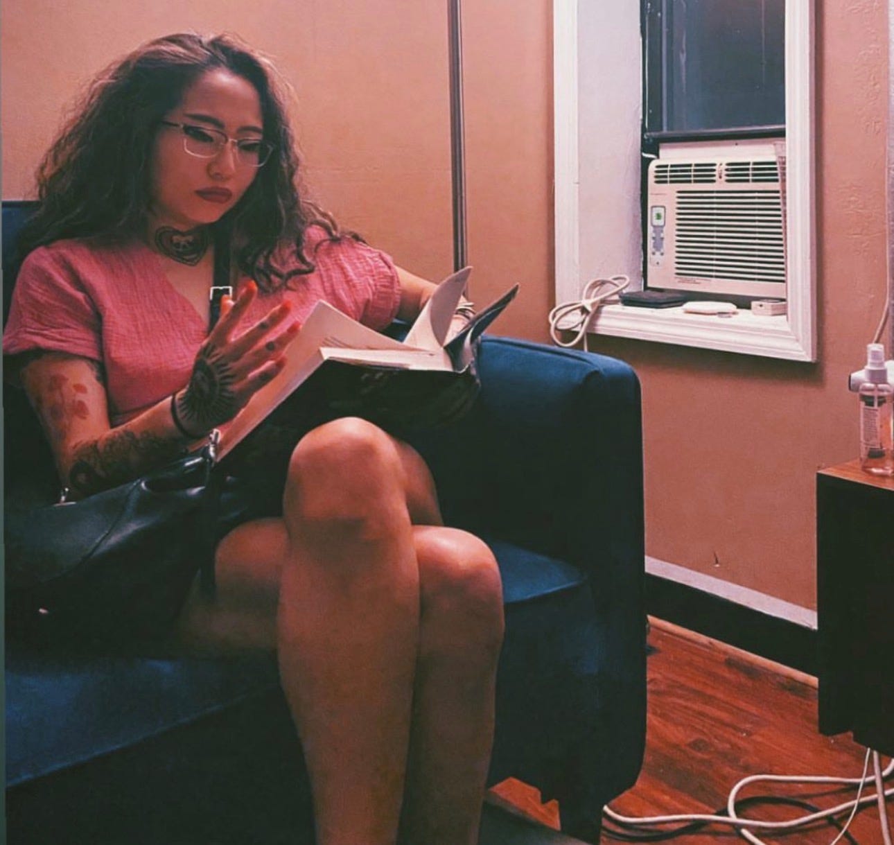 The author, in glasses and a pink top, reads a book cross-legged in a blue armchair beside a window AC unit.
