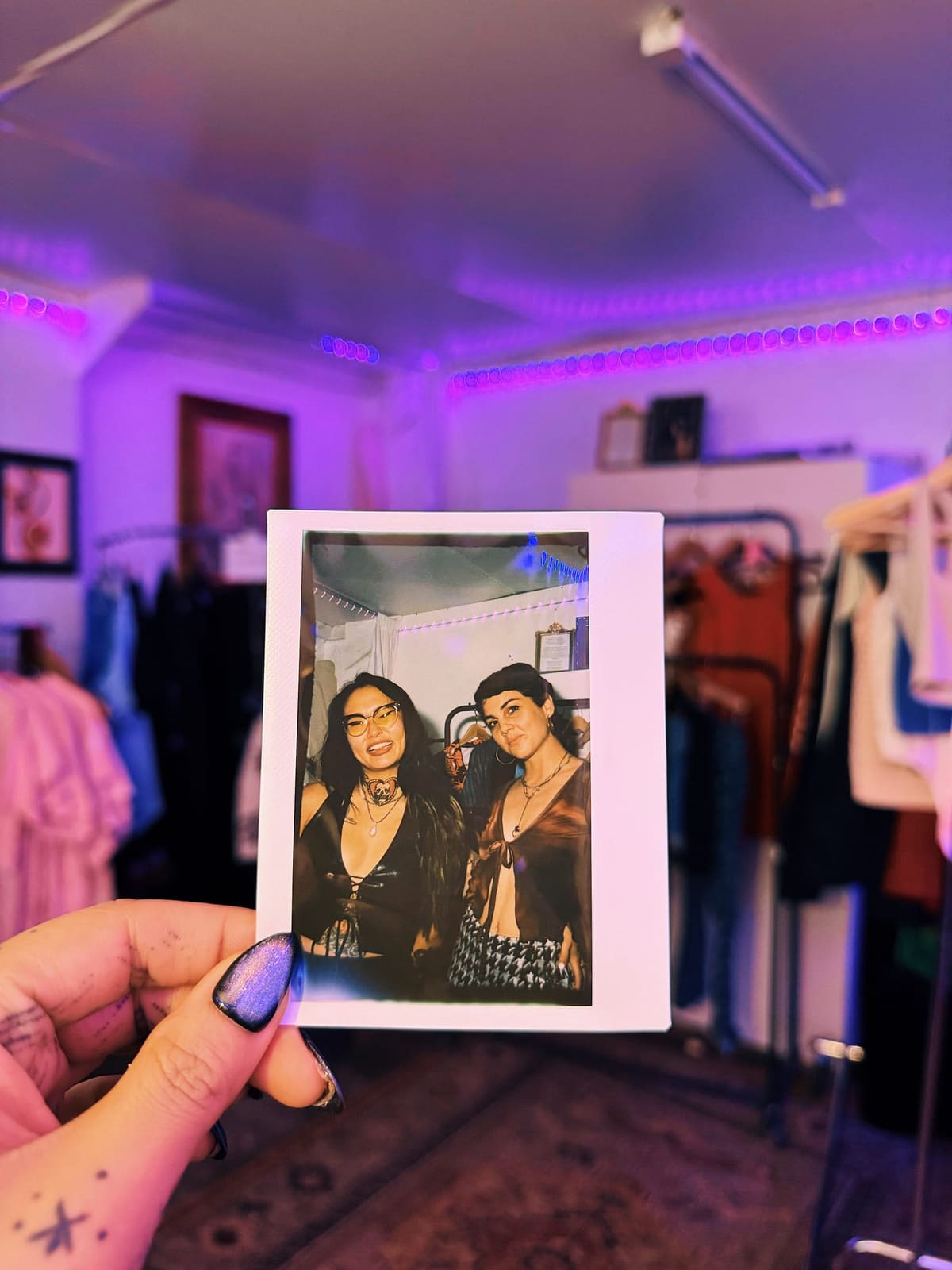 A hand holds a Polaroid of the author and a companion inside a purple-lit pop-up shop lined with clothing racks.