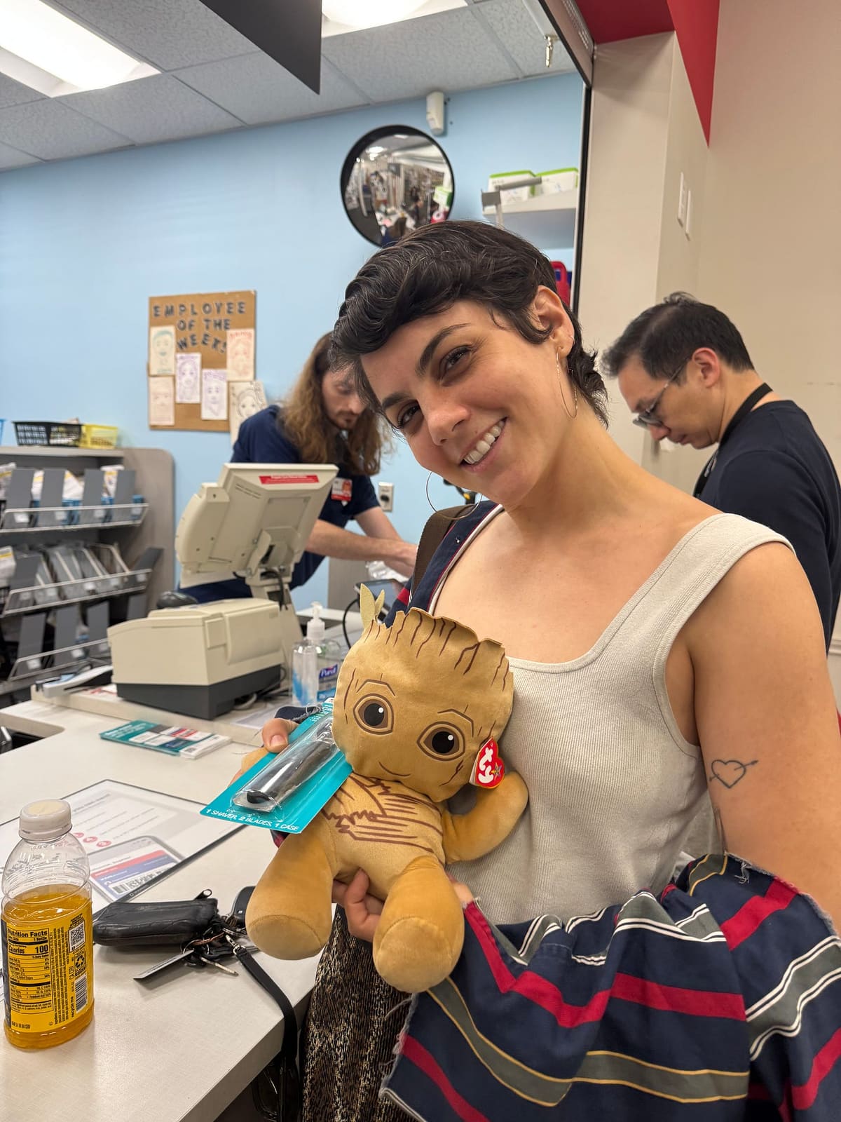 A friend grins at the pharmacy checkout, hugging a plush Groot toy still in its packaging.